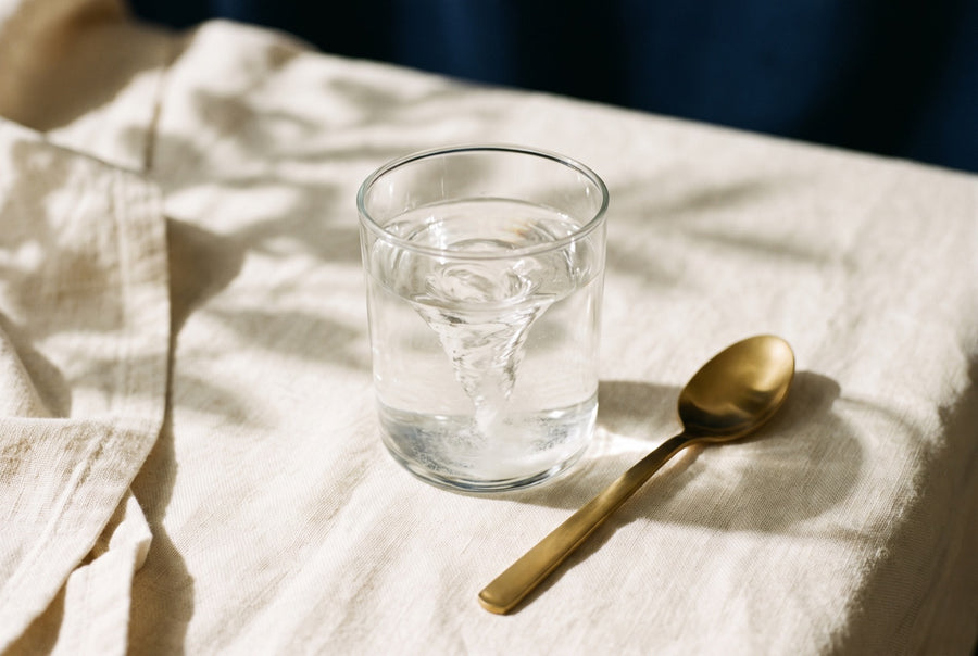 White powder swirling into a vortex in a glass of water, with a gold spoon on cream linen