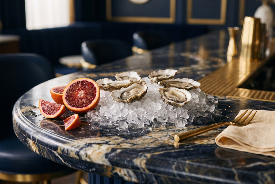 Fresh oysters on crushed ice beside a halved blood orange on a navy marble bar
