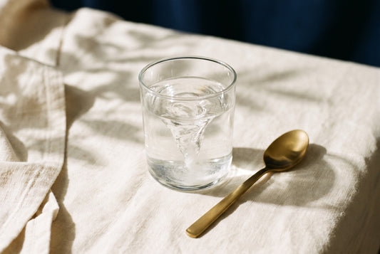 White powder swirling into a vortex in a glass of water, with a gold spoon on cream linen
