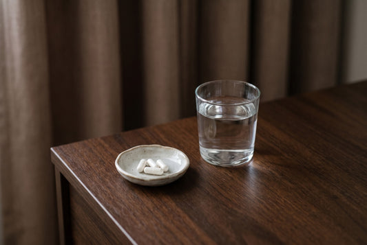 A glass of water and a small dish of white capsules on a bedside table in soft morning light