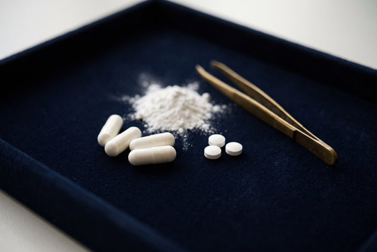 White capsules, small tablets and a heap of powder with brass tweezers on a navy velvet tray